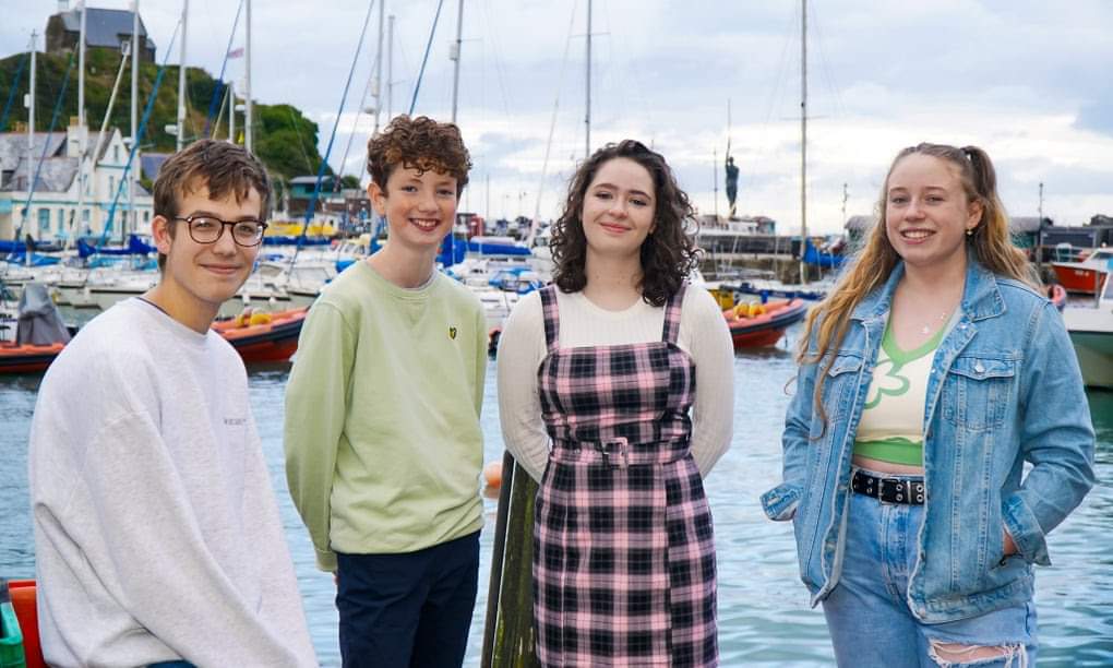 Four teenagers are standing in front of a harbour full of boats. They’re smiling and they look really friendly.
