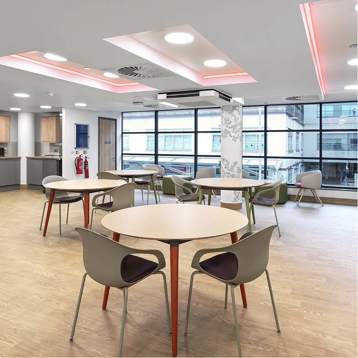 Modern, bright communal area with round tables and comfortable chairs, featuring large windows and contemporary lighting.
