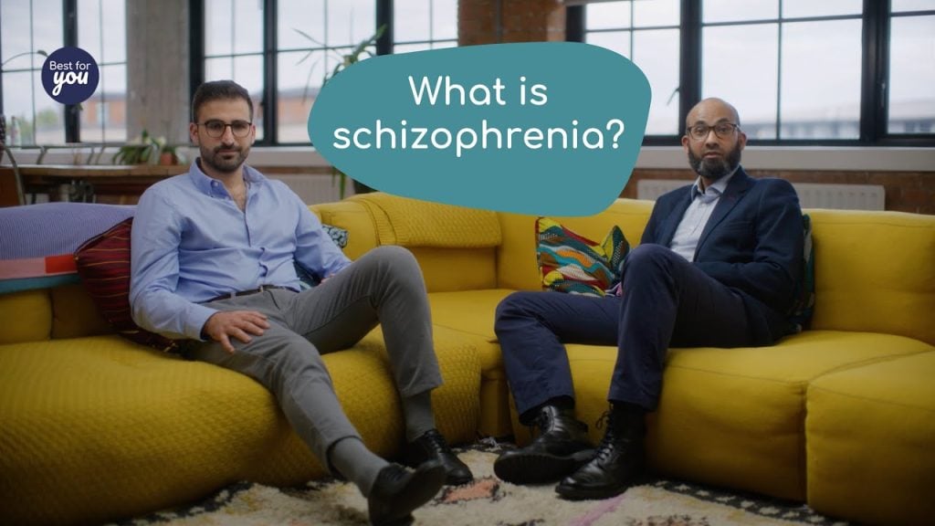 A detailed description of two men sitting on a yellow sofa in a modern office setting, with a speech bubble reading "What is schizophrenia?".