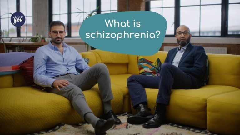 A detailed description of two men sitting on a yellow sofa in a modern office setting, with a speech bubble reading "What is schizophrenia?".